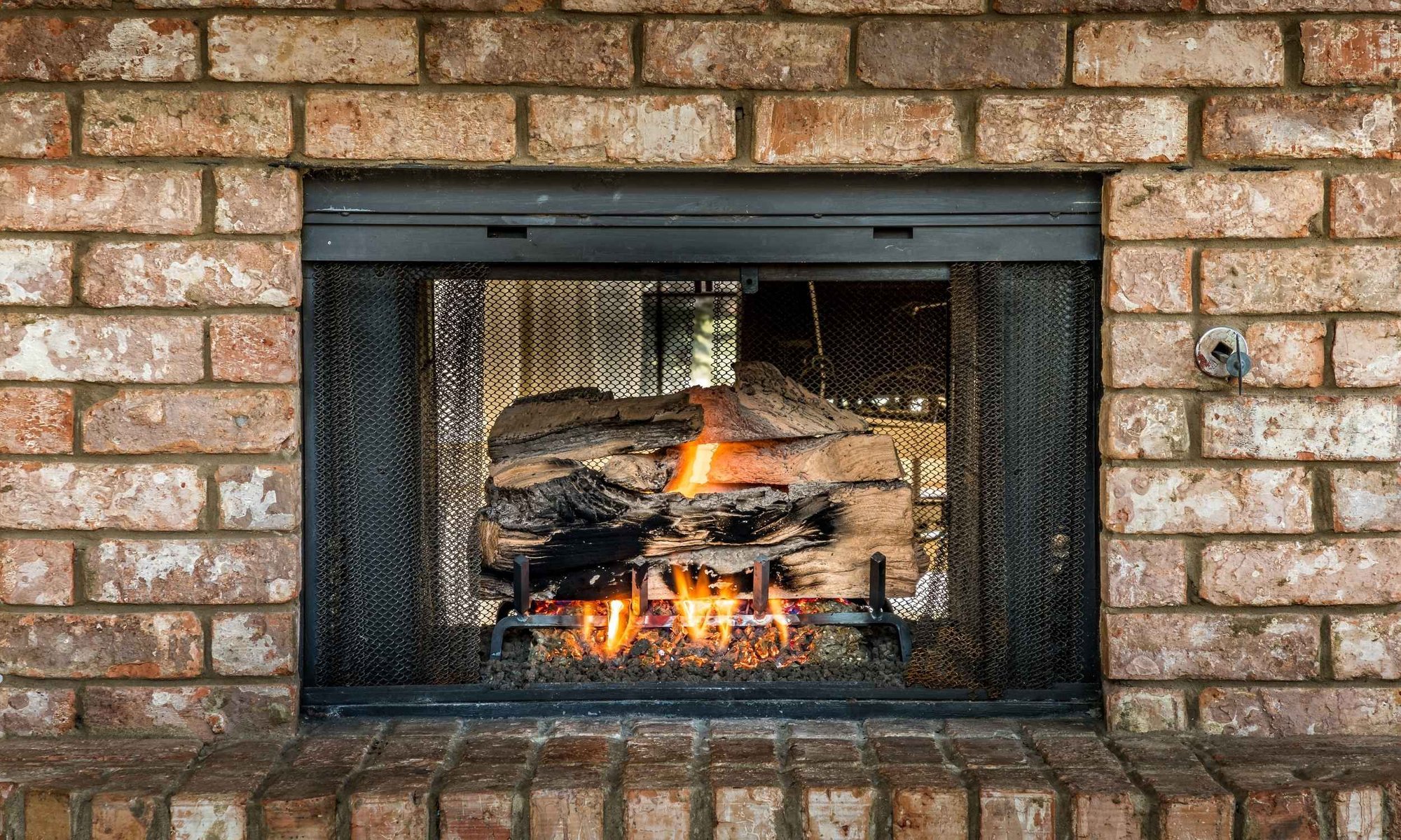 Close-up of a gas log fireplace burning inside a brick fireplace with a black mesh safety screen and hearth.
